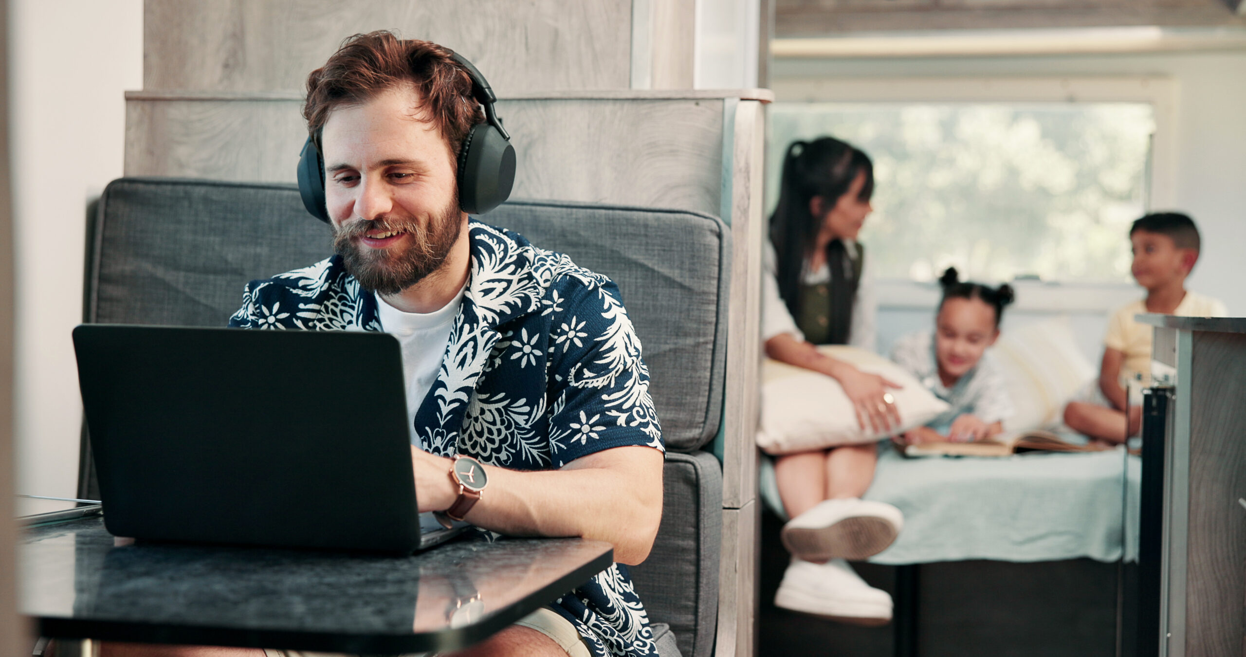 Man with headphones working on a laptop in the lounge area of a modern RV while family relaxes in the background