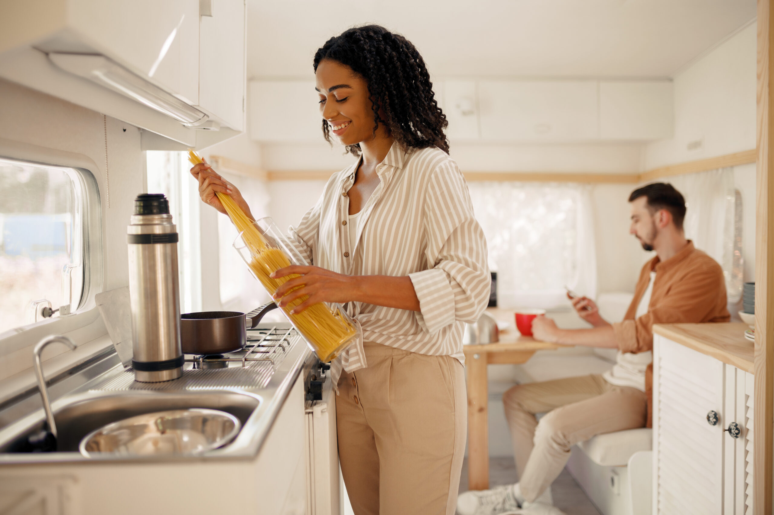 Smiling woman preparing pasta in a bright white RV kitchen while a man sits at the dinette in the background