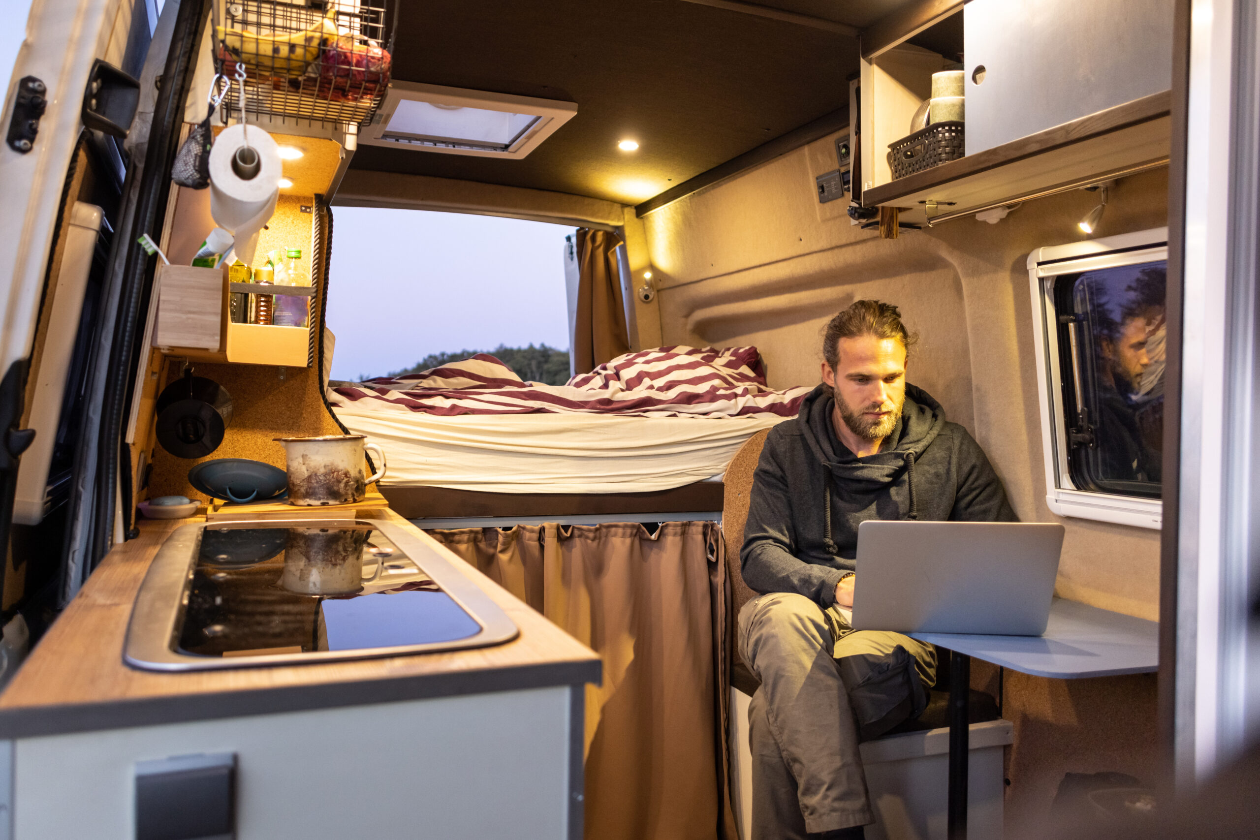 Man with long hair working on a laptop inside a wood-paneled camper van with a bed and small kitchen visible